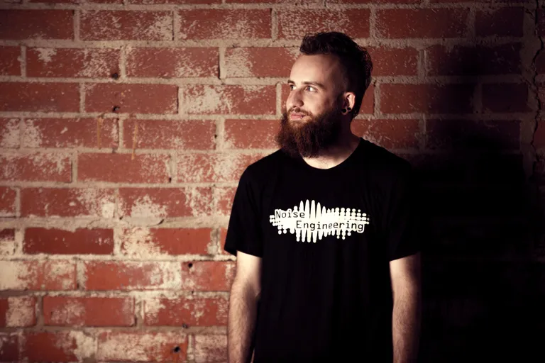 An incredibly attractive (dare I say, gorgeous) person standing in front of a brick wall wearing a t-shirt with the Noise Engineering logo on the front. 