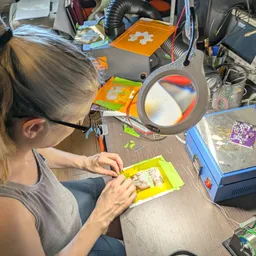 Kris at her desk with a hotplate, a magnifying glass, and a PCB with a yellow stencil covering it